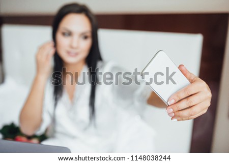 The young beautiful brunette happy woman lies in her bed in the bed with a laptop and a large bunch of roses, the girl makes selfie on a white phone. Close up.