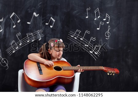 Schoolgirl sitting in front of a blackboard in the classroom and playing the guitar