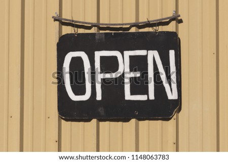 Open signboard with white painted letters on a black weathered board against a yellow wall.