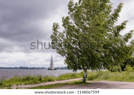 A lonely tree on the river bank and a old flooded chapel beyond on a gray clouds background