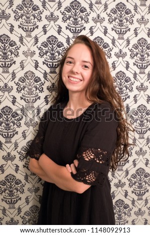 Portrait of a young woman standing in front of classic 1960s vintage fuzz on foil wallpaper. Portrait of brunette with long hair wearing a black dress with arms folded.
