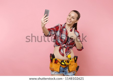 Strong excited young handyman woman in plaid shirt, denim shorts, kit tools belt full of instruments doing selfie on mobile phone isolated on pink background. Female in male work. Renovation concept