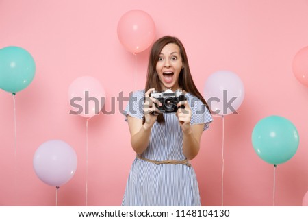 Portrait of amazed young happy woman wearing blue dress holding retro vintage photo camera on bright pink background with colorful air balloons. Birthday holiday party people sincere emotions concept