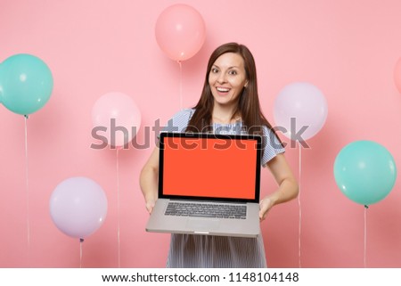 Portrait of amazed happy young beautiful woman in blue dress holding laptop pc computer with blank empty screen on pastel pink background with colorful air balloons. Birthday holiday party concept