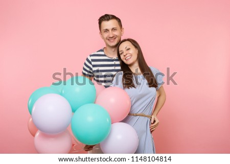 Portrait of young happy smiling couple in love. Woman and man in blue clothes celebrating birthday holiday party on pastel pink background with colorful air balloons. People sincere emotions concept