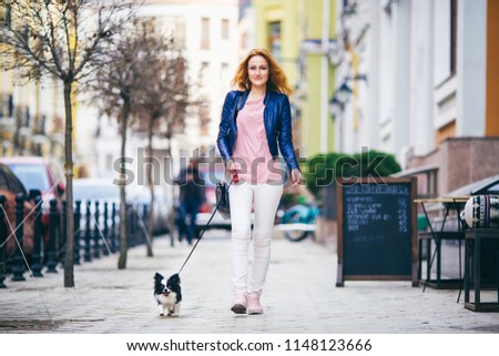 A young redhaired Caucasian woman walking along European street with small Chihuahua breed dog of two colors on leash. Cloudy, warm autumn spring weather. Girl Dressed in leather jacket and pink shoes
