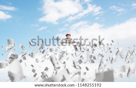 Young little boy keeping eyes closed and looking concentrated while meditating among flying papers in the air with cloudy skyscape on background.