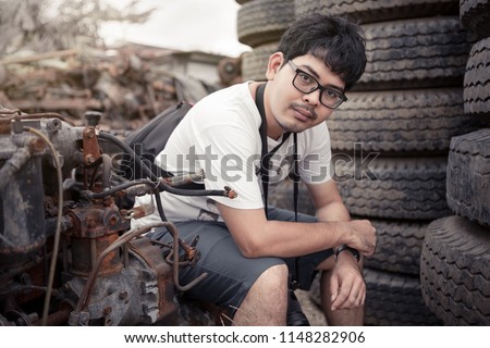man with glasses in metal scrap junk yard surrounding with vehicle and engine parts, tires and metal scrap. He hold DSLR camera like foreigner tourist.
