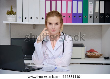portrait of a beautiful blonde girl doctor sitting at a table in the office with a laptop. She is right in front of the camera smiling and looks serious