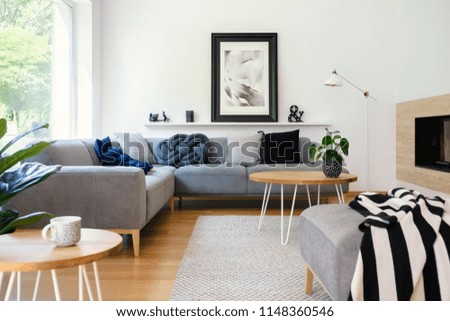 Poster on white wall in living room interior with table in front of corner sofa with pillows. Real photo