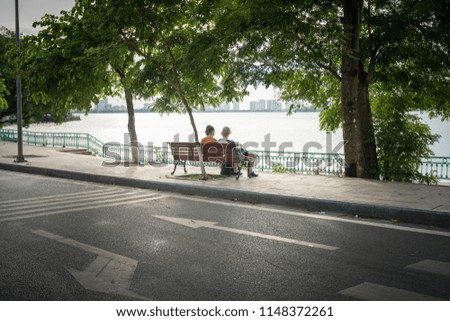 Two young women sitting on bench under big tree by the lake in city