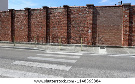 Urban background. Street with crosswalk in front of a concrete sidewalk, yellow metal bollards and an old brick wall in a bright sunny day