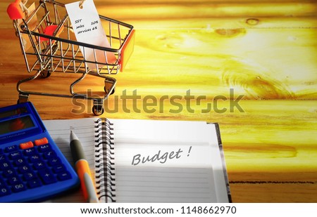 Conceptual of Sales Services Tax in Malaysia starting Sept'18. Dark texts on white tag on mini trolley chart and blank note book on wooden table.  Focus on trolley. Pen, calculator, others in blur.