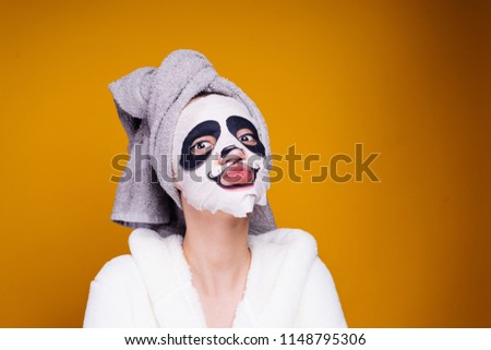 a funny young girl with a towel on her head and a white robe takes care of herself, on her face a mask with a panda face