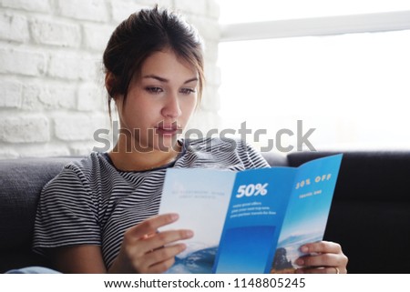 Young hispanic woman laying on couch at home, holding a travel flyer and planning next trip. Copy space on window. Royalty-Free Stock Photo #1148805245