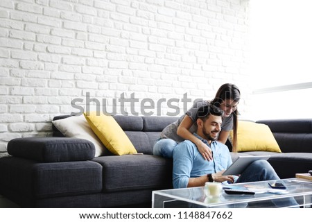 Young hispanic couple sitting on couch at home, using a tablet PC for Internet and social media. The girl is giving a massage to her boyfriend. Copy space Royalty-Free Stock Photo #1148847662