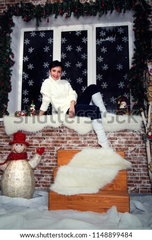 brunette girl in white sweater and white Golf in jeans sitting on a window with snowflakes wood and snowman concept new year in rustic style