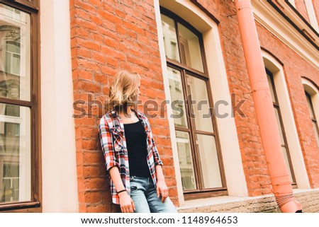 A beautiful and young girl stands near a brick house with large wooden windows. Her hair is loose and her head is turned to the left so that her face can not be seen.