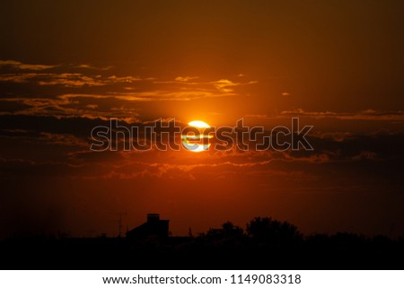 Sunset with strong orange colors with clouds in front of the sun and the dark silhouette of Olching, Germany in the bottom of the picture