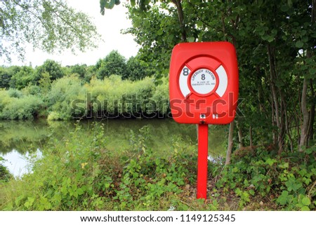 Wide shot of a red Lifebuoy Station atop its metal pole surrounded by trees & vegetation overlooking a river ready to be used by the public or emergency services for swimming or boating emergencies.