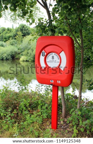 Angled shot of a red Lifebuoy Station atop its metal pole surrounded by trees & vegetation overlooking a river ready to be used by the public or emergency services for swimming or boating emergencies.