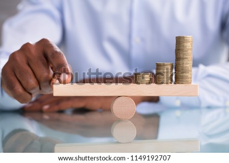 Close-up Of A Businessman Balancing The Coin Stack On Wooden Seesaw Over The Reflective Desk Royalty-Free Stock Photo #1149192707