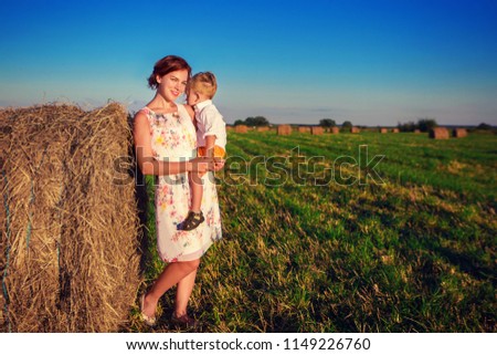 A woman is holding a baby in her arms. Mom and son are standing near a haystack in the field