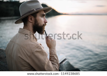 Portrait of young bearded writer writes notes in a notebook, enjoying beautiful nature and sunset, planning travel.  Handsome traveller wearing hipster hat sitting on the river bank and thinking.  Royalty-Free Stock Photo #1149238685
