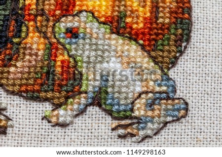 Cross-stitch embroidery with cat in hat, cauldron, toad, bonfire and pumpkin.Part art on mouline threads with grey frog and yellow pumpkin. Close-up.