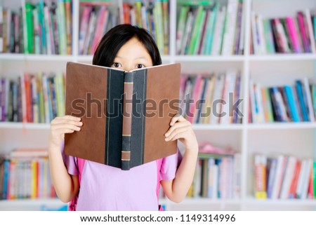 Picture of a cute schoolgirl looking at the camera and covering her face with a book while standing in the library
