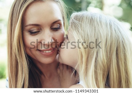 Waist up portrait of smiling female sitting outside. Little kid is kissing her cheek with care and tenderness. They are pleased and positive