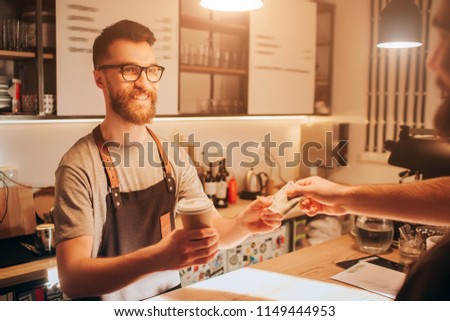 A picture of barman standing behind the bar stand and holding a cup of coffe that he did for the customeer. The barman looks happy and smiling while getting a cash for order from customer.
