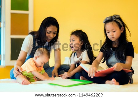 Babysitting playing with three foolish playful kids carrying on the bed; widow young woman and child are enjoying funny time indoor in modern yellow bed room.
