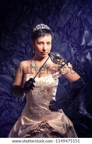 brunette girl with short hair in crown with rhinestones and choker in ballroom, wedding dress with black satin gloves and black cloak with masquerade lace mask for new year on black uneven background
