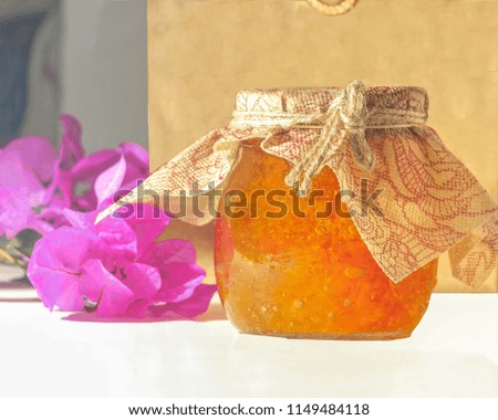 Orange  homemade marmalade in a glass jar . Pink flowers in the background with a brown paper bag. Stock Image.
