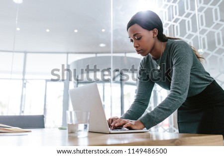 Young woman standing by conference table and working on laptop. Female preparing a business proposal before a meeting in boardroom. Royalty-Free Stock Photo #1149486500