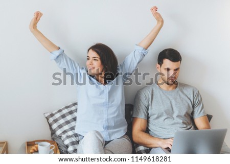 Young married couple spend morning in bed: beautiful brunette female stretches and her husband with serious expression uses laptop computer for remote work, dressed casually, being in bedroom