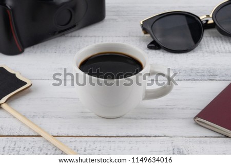 A cup of coffee, passport, sunglasses, film camera and blank small blackboard on white wood background, selective focus on coffee