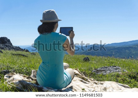 girl in dress and hat sitting on a blanket with a tablet in her hands on an alpine meadow against a backdrop of mountains, the view from the back