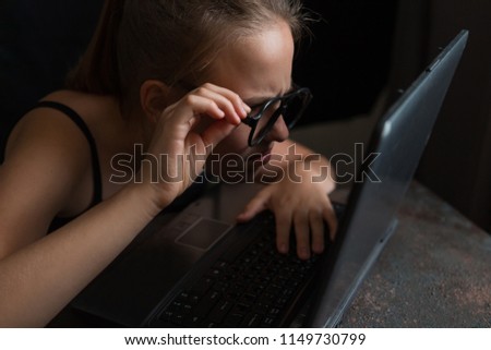 teenage girl in glasses works at a computer at night and peers at a monitor on a dark background with a copy of space, the concept of freelancing and vision problems