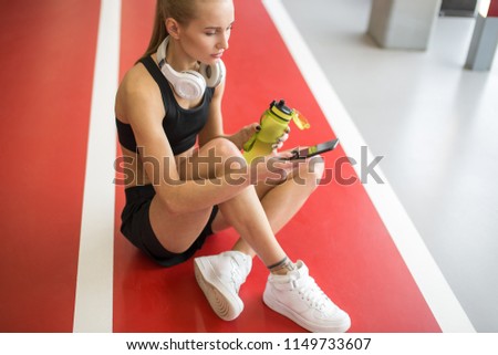 Young blond woman with mobile phone sitting on floor and holding water bottle, chatting in smartphone
