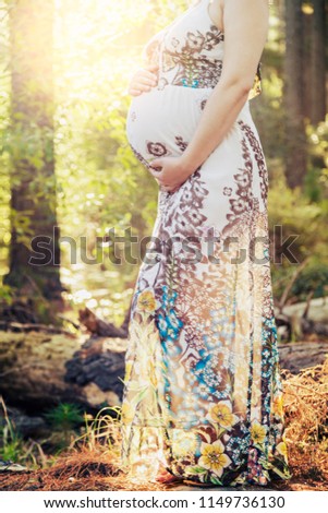 A cropped look at a young woman standing in a sunlit forest; she is very pregnant and cradles her rounded belly gently.