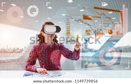 Young woman in checkered shirt using VR headset with digital media interface while sitting inside bright office building. Virtual reality technologies for education
