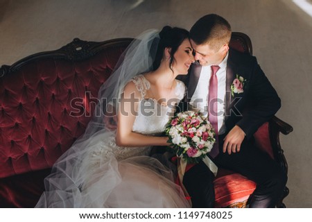 Beautiful newlyweds are sitting in the studio on a red armchair under a sun ray. A gentle wedding portrait of a young groom and a cute bride in a stylish studio.