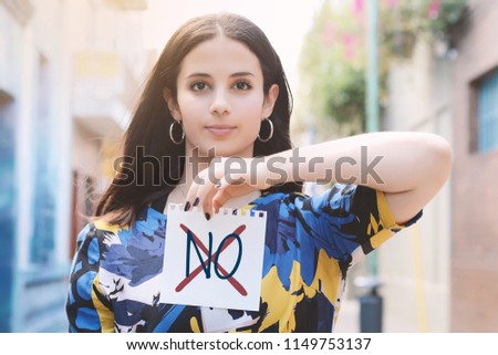 Closeup of a young woman outdoors showing a notepad with the text "no" written in it.