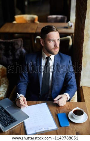 Serious pensive handsome young man in formal suit sitting in armchair and looking out window while thinking about solution in business cafe