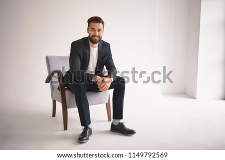 People, lifestyle, business, style, fashion and men's wear concept. Positive successful young CEO sitting in armchair, smiling at camera, dressed in elegant shoes, trousers, jacket and white t-shirt Royalty-Free Stock Photo #1149792569