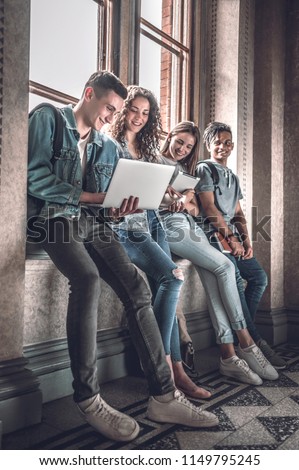 Confident students.Group of happy young people working together and looking at laptop while sitting at the window sill indoors. Royalty-Free Stock Photo #1149795245
