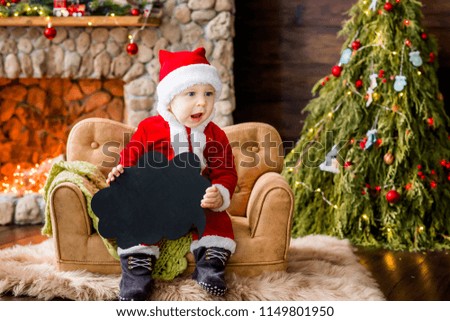 A small child in a red Santa Claus costume, sitting on the couch in the residence, holds a black cloud plate for text in his hands. New Year card. The blond boy laughs. Christmas tree with boxes 