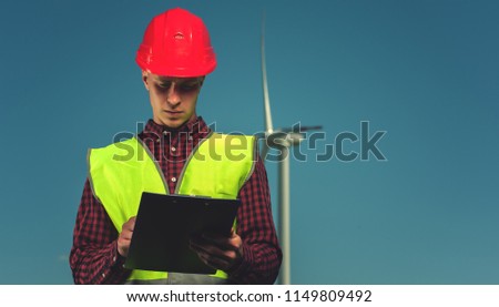 Young handsome male chief engineer in a green waistcoat and red protective helmet signs a design of engineering works against the backdrop of a windmill and a blue sky.